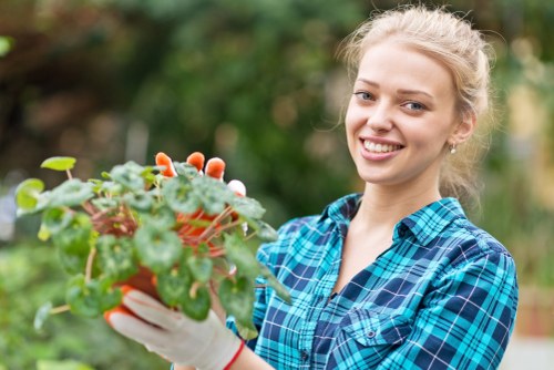 A young woman with blonde hair tied back, wearing a blue and green plaid shirt and gardening gloves, is standing outdoors in a garden. She is smiling and holding a small potted plant with green foliage and reddish leaves, positioned in front of a background featuring lush greenery, including blurred trees and shrubs. The garden setting has a natural, well-maintained appearance, with various plant elements visible, suggesting it is part of a landscaped outdoor space in the Kings Cross area. The lighting indicates a bright, possibly sunny day, emphasizing the vibrant green tones and creating a warm, inviting atmosphere suitable for gardening and outdoor maintenance services.
