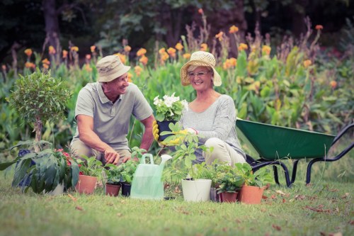 A mature couple is sitting in a well-maintained garden with vibrant flowering plants and lush green foliage, surrounded by a variety of potted plants and garden tools. The man and woman are wearing light-colored, wide-brimmed hats suitable for sunny weather, and they are smiling while engaging in gardening activities. The woman holds a white flowering plant, while the man appears to be tending to other plants nearby. Behind them, tall purple flowers and a mix of green shrubs create a natural, colorful backdrop, indicating a thoughtfully landscaped outdoor space. The garden features a neatly mown lawn, a green wheelbarrow, and gardening containers, emphasizing outdoor maintenance and care typical of professional garden landscaping services in Kings Cross or surrounding areas. The scene is set under bright weather conditions, with natural sunlight illuminating the scene, capturing an inviting and lush garden environment that aligns with high-quality gardening and landscaping services offered by Landscapers Kings Cross.