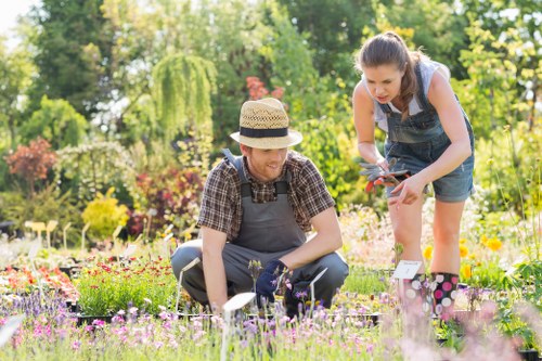 A man and a woman gardening together in a well-maintained outdoor garden, with lush green trees, flowering plants, and neatly arranged flower beds in the background. The man, wearing a straw hat, checkered shirt, and gardening gloves, is kneeling on a paved pathway and tending to small flowering plants in a flower bed. The woman, dressed in a sleeveless top and denim shorts, is standing nearby, holding gardening shears and assisting with plant maintenance. The garden features a mix of vibrant blooms in various colours, including pinks, purples, and yellows, with a tidy lawn and paved walkways creating a structured outdoor space. Bright natural sunlight illuminates the scene, suggesting clear weather. The setting appears to be a suburban or residential garden in Kings Cross or a nearby area, with a focus on gardening activities that thrive with professional landscape and garden maintenance services, such as those provided by landscaperskingscross.co.uk, emphasizing outdoor care and enhancement.