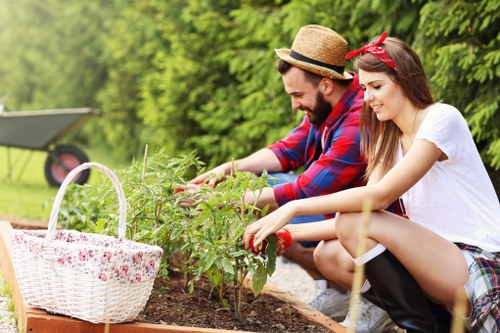 A man and a woman are working together in a neatly maintained garden, planting or tending a small shrub within a raised flower bed. The garden features a lush green backdrop with mature trees and a well-kept lawn. The woman, wearing a white T-shirt, patterned shorts, and a red headband, is smiling as she handles the plant. The man, dressed in a red plaid shirt and a straw hat, is also engaged in the gardening activity. Nearby, on the left side of the image, there is a white wicker basket with a floral pattern placed on the edge of the flower bed, possibly containing gardening tools or supplies. The garden appears to be part of a residential outdoor space, with natural sunlight illuminating the scene and a slightly blurred background showing a garden shed or equipment. This setting exemplifies typical garden maintenance and planting activities supported by professional gardening services such as those provided by Landscapers Kings Cross, which may include planting, soil preparation, and garden care in the Kings Cross area of London.