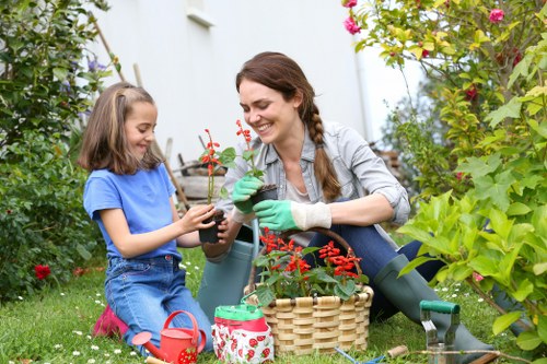 A woman and a young girl are gardening together in a lush outdoor garden space, surrounded by various green plants, shrubs, and flowering bushes. The woman, wearing a light grey jacket and green gardening gloves, is smiling as she helps the girl hold a potted flowering plant, possibly a small red-flowered shrub. The girl, dressed in a blue t-shirt and jeans, appears engaged and happy while participating in planting activities. A wicker basket, small gardening tools, and a decorative watering can rest on the grass nearby, indicating ongoing gardening work. The garden features a mix of dense foliage, colorful blooms, and well-maintained grass, with natural daylight illuminating the scene, suggesting a bright, pleasant day suitable for outdoor lawn and garden care typical of landscaping projects in the Kings Cross area. This image reflects active outdoor maintenance and gardening activities associated with professional services offered by Landscapers Kings Cross.