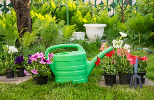 The image shows a well-maintained garden area with lush green grass in the foreground, featuring a large, bright green plastic watering can placed among potted flowering plants. These potted plants display vibrant blooms in shades of pink, white, and purple, arranged along the garden's edge. Behind the plants, there is a backdrop of various shrubs, ferns, and a mature tree with a textured trunk, providing shade and structure to the outdoor space. In the background, a white decorative garden urn is positioned near dense greenery, including trees and leafy bushes, creating a natural, somewhat shaded environment that suggests a landscaped backyard or front garden. The scene appears to be taken during mild or partly cloudy weather, with natural daylight illuminating the scene evenly, highlighting the fresh and healthy appearance of the plants and the neatness of the lawn and garden features. The overall setting reflects the outdoor spaces where professional gardening and landscaping services, such as those offered by Landscapers Kings Cross, support garden maintenance and enhancement efforts in the local area of Kings Cross, London, N1 postcode.