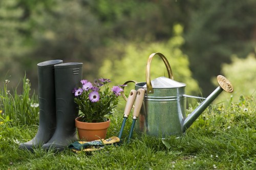 A neatly arranged collection of gardening tools and supplies placed on a lush green lawn in a garden setting. The foreground features a pair of black rubber gardening boots standing upright beside a small terracotta pot filled with purple and white flowering plants. Nearby, various hand tools such as a trowel, pruning shears, and a small fork are visible, leaning against a silver watering can with a wooden handle. The garden surface is composed of well-maintained grass, with a backdrop of blurred trees and foliage indicating a mature, landscaped outdoor space in a residential area near Kings Cross, London. The natural lighting suggests a bright, partly cloudy day, ideal for outdoor gardening work, which aligns with the services offered by Landscapers Kings Cross in maintaining and enhancing garden landscapes across the area.