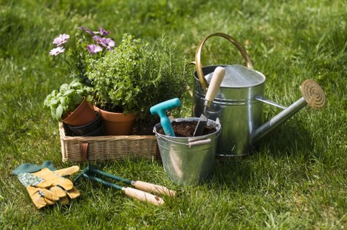 In a well-maintained outdoor garden area, a collection of gardening tools and supplies are arranged on a lush, green lawn. The scene features a rectangular wicker planter filled with various potted plants, including leafy greens and flowering blooms in shades of purple and white. Nearby, a pair of yellow gardening gloves rest on the grass, alongside small hand tools such as a trowel and pruning shears with green handles. To the right, a metallic watering can with a wooden handle and a silver spout is positioned next to a metal bucket containing gardening implements. The surrounding grass is dense and evenly clipped, with a rich, natural green tone, suggesting recent care or ongoing gardening activities. The background shows an outdoor environment typical of a suburban garden in Kings Cross, with natural lighting indicating a clear day. The arrangement of tools and plants reflects a gardening project in progress, supported by local landscapers, with a focus on outdoor plant care and lawn maintenance.