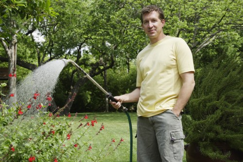A man standing in a lush, well-maintained garden in Kings Cross, London, holding a hose and watering vibrant red and green flowering plants. The garden features a neatly trimmed, lush green lawn in the foreground, bordered by flower beds with blooming plants. Behind him, there are mature trees with dense foliage, creating a shaded outdoor environment. To the right, a dense hedge and bushy shrubs add privacy and structure to the landscaped yard. The weather appears bright and clear, with natural sunlight illuminating the scene, emphasizing the healthy growth of the garden elements. This outdoor space reflects professional gardening and landscape maintenance, possibly serviced by Landscapers Kings Cross, aligning with their comprehensive planting and garden care services.