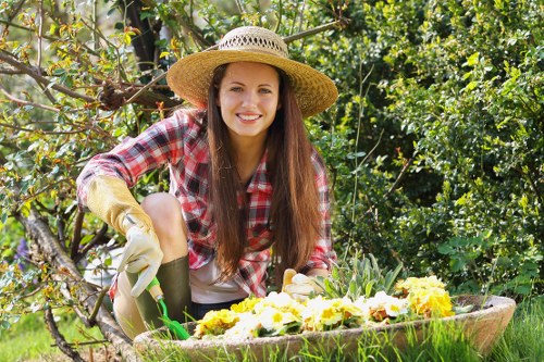 A young woman with long brown hair, wearing a wide-brimmed straw hat, a red plaid shirt, and gardening gloves, is kneeling in a lush garden with vibrant green foliage and flowering plants. She is smiling at the camera while tending to a wooden planting box filled with yellow and purple flowers, which are arranged along the edge of the garden bed. Behind her, there is a small tree with twisting branches and a mix of leafy shrubs, indicating a well-maintained outdoor space suitable for gardening activities. The scene is brightly lit, suggesting a sunny day, and the garden features a combination of neatly trimmed bushes, flower beds, and natural soil patches. This setting captures an example of sustainable outdoor gardening practices, aligning with services offered by Landscapers Kings Cross in the London area, contributing to environmentally conscious landscaping and garden management. The overall environment reflects a thriving, organized garden space designed for both visual appeal and ecological sustainability, with attention to plant selection and landscape structure typical of professional gardening in central London surroundings.