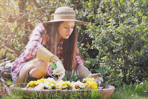 In a well-maintained outdoor garden area, a young woman wearing a wide-brimmed straw hat, a plaid shirt, and gardening gloves is kneeling on the grass while tending to a colorful flower bed. The flower bed is bordered by a low, natural stone edging and contains a variety of seasonal blooms in shades of yellow, white, and purple. Behind her, a lush hedge and mature trees provide a green backdrop, with sunlight filtering through the leaves, casting dappled light across the scene. The grass in the foreground is dense and healthy, contrasting with the soil in the flower bed. The overall scene depicts a peaceful, landscaped outdoor space suitable for gardening and lawn care, reflecting professional landscaping standards that a company like Landscapers Kings Cross might implement in the local area near Kings Cross, London, with visible tools and cultivated plantings indicative of thorough garden maintenance and restoration work.