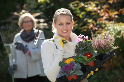 A young woman with blonde hair, wearing a cream sweater and yellow gardening gloves, is smiling and holding a black plastic tray filled with various potted flowering plants, including purple, yellow, and red blooms, in a well-maintained garden with green grass, shrubs, and blooming flowers in the background. An older woman with glasses, wearing a white jacket and a dark scarf, stands behind her holding a garden rake. The garden scene suggests outdoor maintenance or planting activities in a landscaped yard, with natural sunlight casting a warm glow on the scene. The setting is indicative of a property that benefits from professional gardening and landscaping services, such as those offered by Landscapers Kings Cross, to maintain vibrant outdoor spaces in the local area, possibly near Kings Cross.